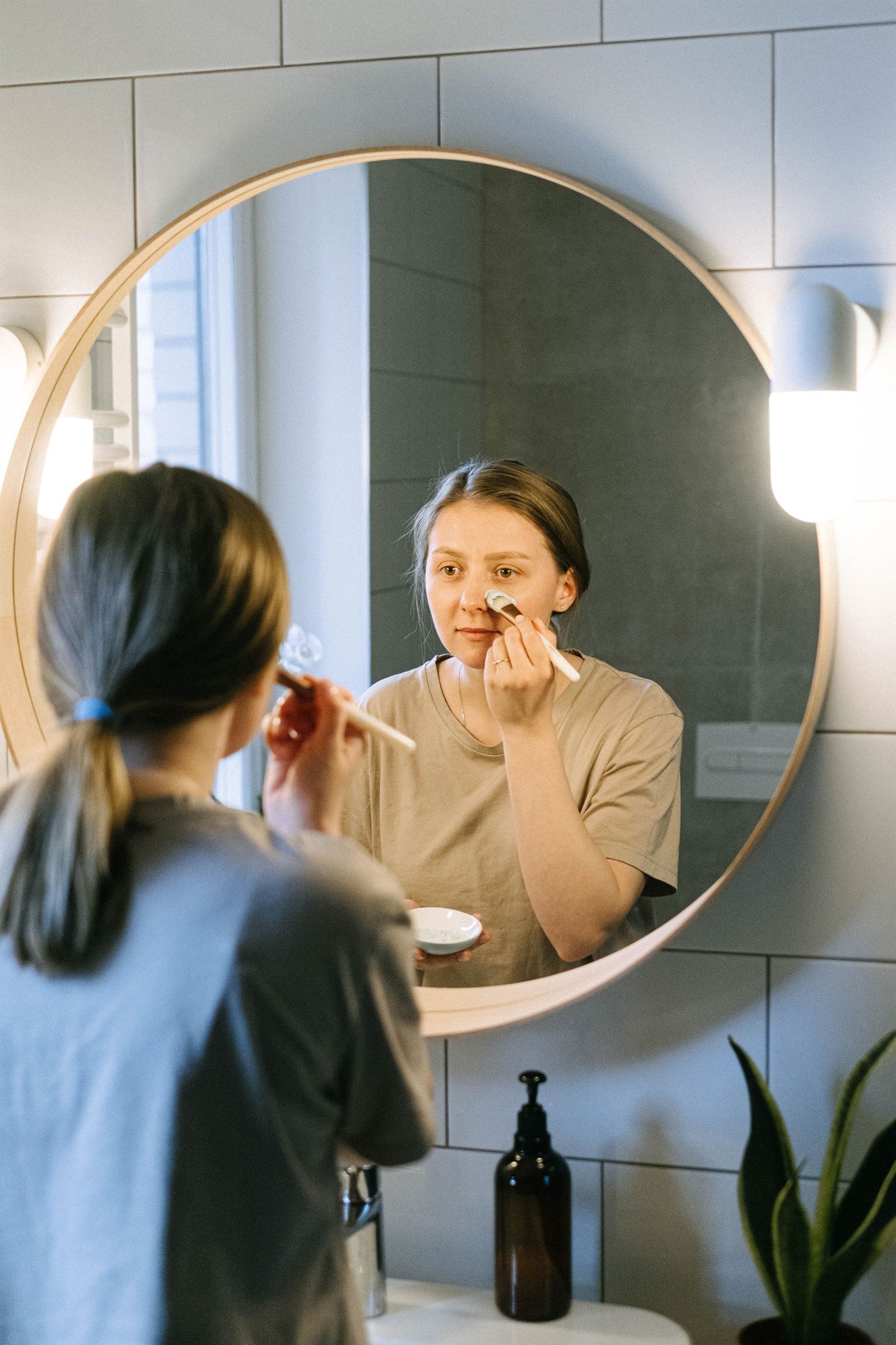 Young woman applying skincare routine at home in front of a bathroom mirror.